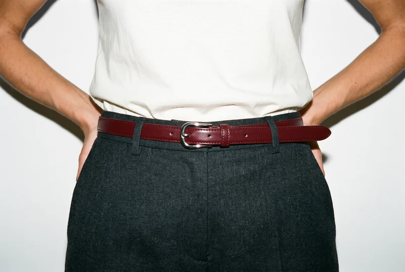 A woman wears an oxblood leather dress belt around her waist, shown from the front with her hands resting on her waist. The belt is framed against a pure white studio backdrop.
