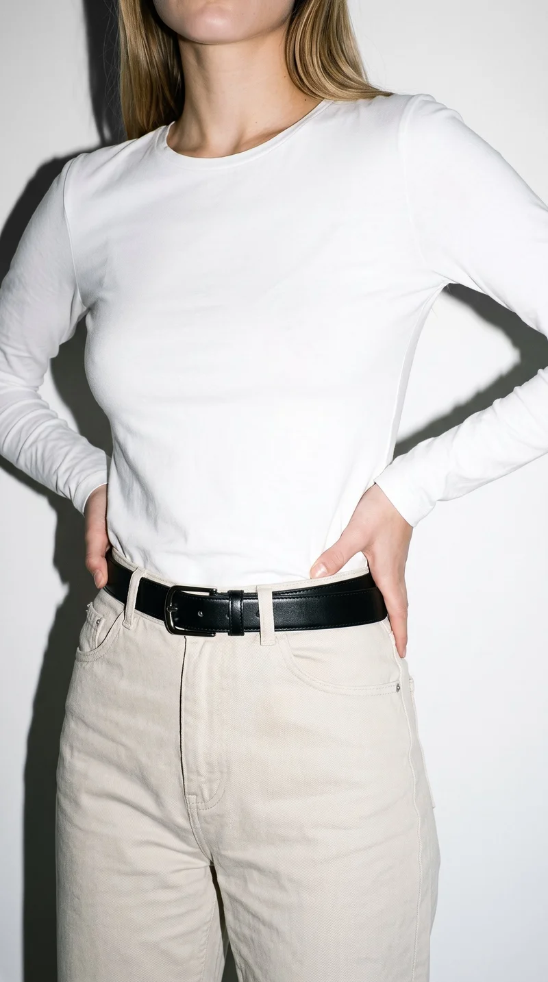 A female model is shown from the torso in an angled front view, wearing a black formal belt with her hands on her hips. The belt is set against a pure white studio backdrop.