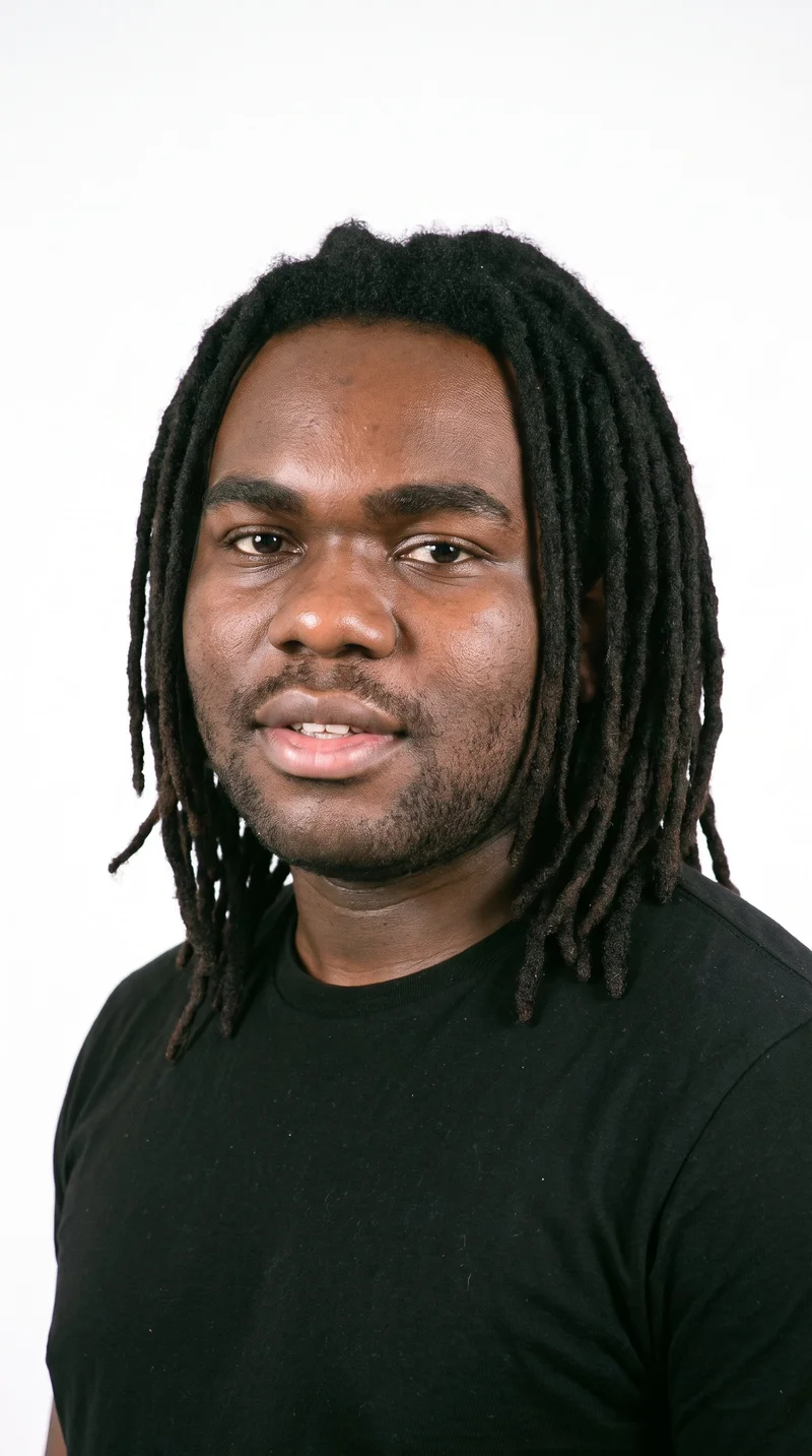This 21-year-old African man has a very dark, deep-brown to black complexion with a cool undertone. His Roman face has strong classical features, a prominent nose line, and a defined jaw. He has thick eyebrows, brown eyes, and full lips. His dark-brown hair is styled in long, rope-like dreadlocks, and dense heavy stubble forms a dark, defined layer across the beard area.