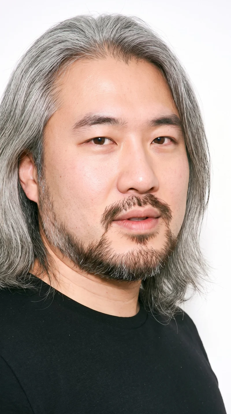 A 38-year-old Chinese man with fair skin and a cool undertone has a square face defined by a broad forehead and strong, angular jaw. He has thick eyebrows, brown eyes, and full lips. His gray hair is long, worn loose past the shoulders with natural volume, and he has a short, full beard trimmed evenly with clean cheek and neckline edges.
