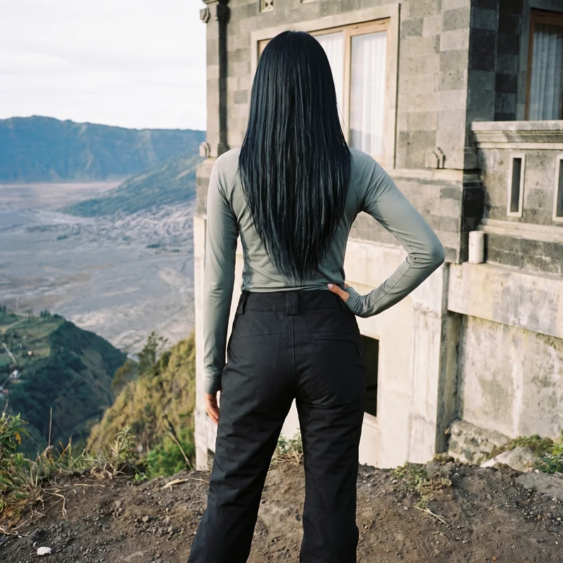 An 18-year-old Southeast Asian woman wears black ski trousers, shown from the back in a three-quarter-length frame with one hand on her waist. A Santorini caldera view forms the backdrop.