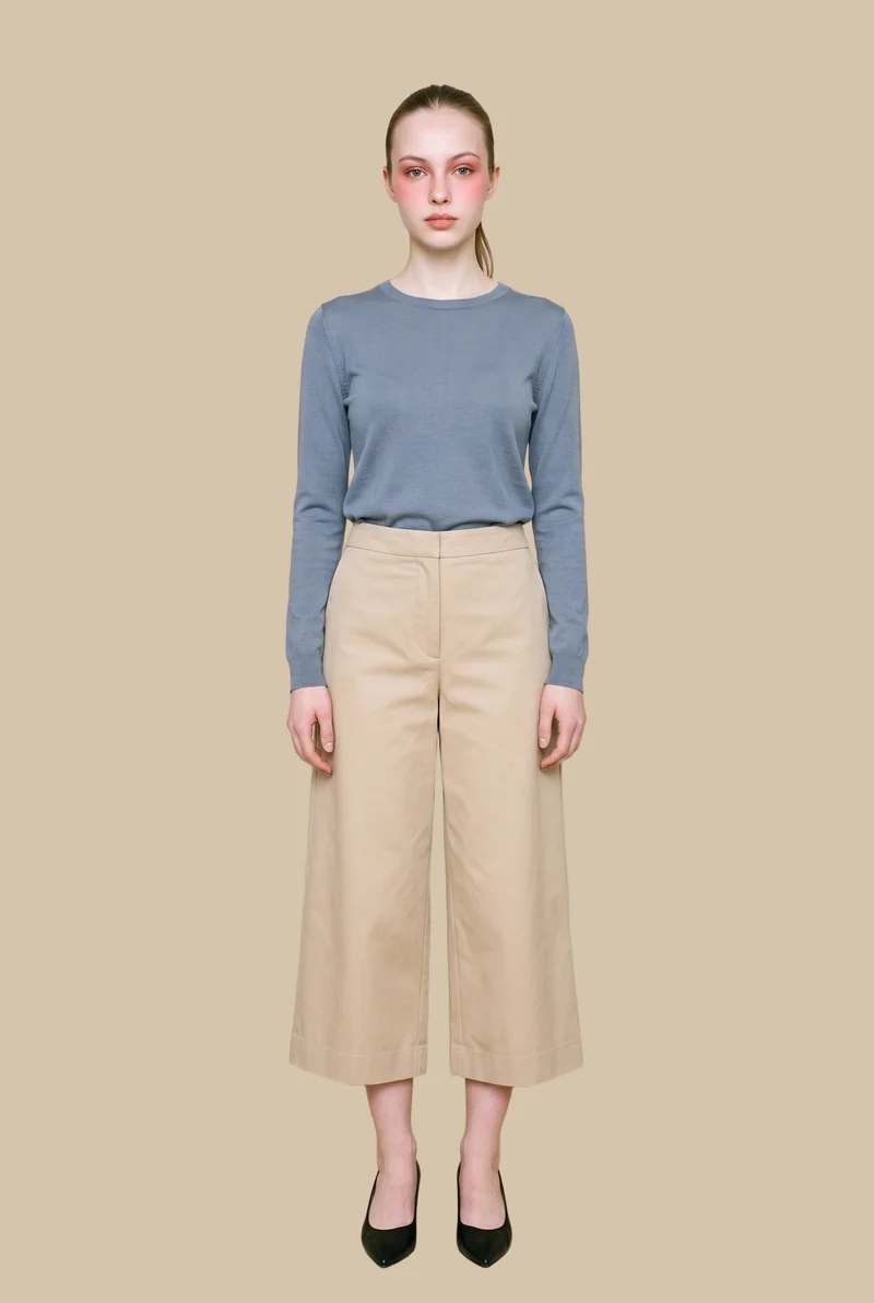 A 21-year-old Caucasian woman wears beige tailored culottes in a full-body front view, standing with one foot forward and a natural expression. A Venetian plaster backdrop fills the background.