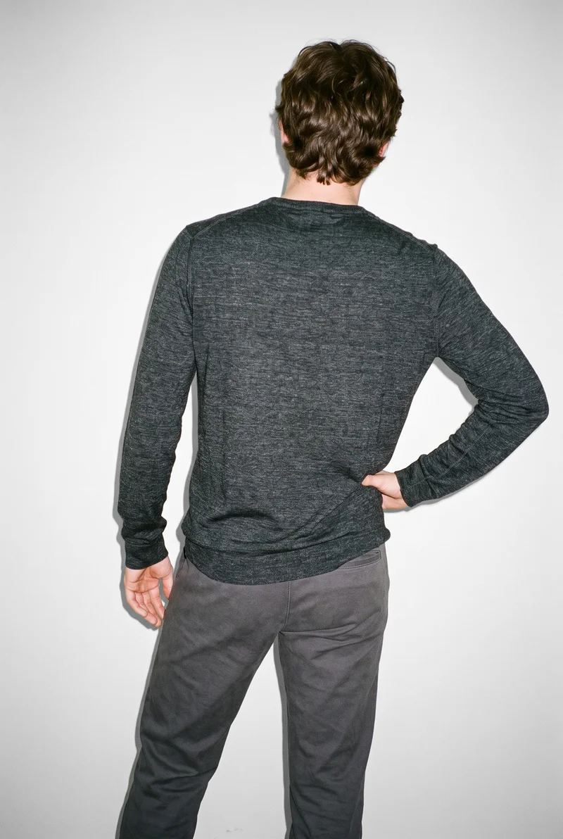 A 21-year-old Caucasian male model wears a grey merino thermal top, shown from the back in a three-quarter-length frame with his weight shifted back. A fog-grey studio wall forms the backdrop.