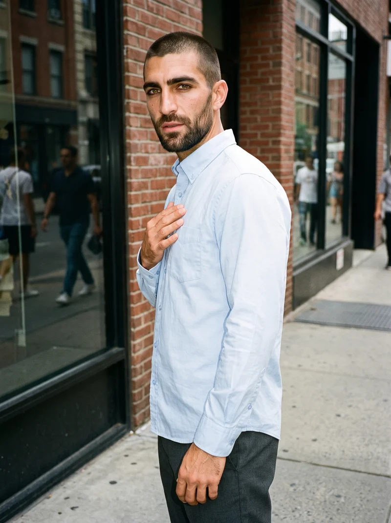 A 33-year-old Caucasian man wears a light blue button-down shirt in an upper-body view. He is shown from a front three-quarter angle with his fingers at his chest and a natural facial expression, against an urban street backdrop.