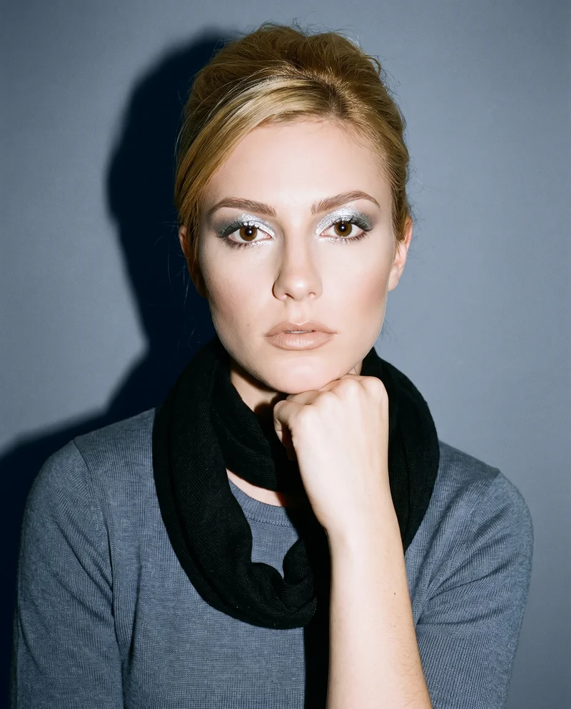 A 21-year-old Caucasian woman wears a black pashmina shawl in a bust-length front view. She rests her chin on her knuckles with a neutral expression against a slate grey studio-wall backdrop.