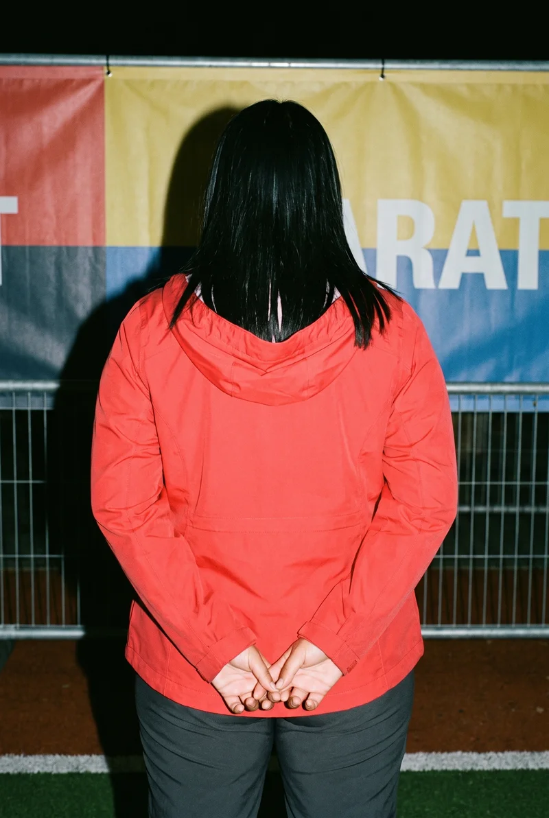 A 22-year-old Arab woman is shown from the upper body in a back view, wearing a red women's hardshell jacket with her hands behind her back. A Marathon Finish Line backdrop is visible.