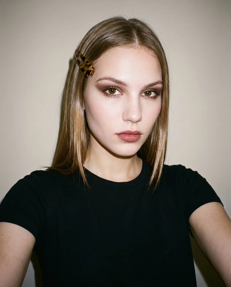 Front-view selfie of a 19-year-old Slavic woman wearing a tortoiseshell hair clip, with her head level and an editorial facial expression. A London Fog studio-wall backdrop frames the composition.