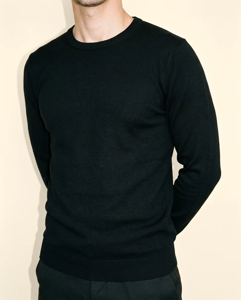 A male model wears a black cashmere knit in an angled front torso view, standing with his hands behind his back. A cream silk studio backdrop fills the background.