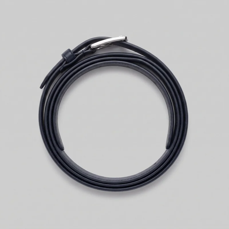 A complete slim navy formal belt is shown as a standalone product, coiled into a circular roll. Its elongated rectangular pin buckle and sliding keeper loop are visible near the top, with a free strap tail extending beside them. Smooth rounded edges, a subtly raised edge profile, and fine stitching along the inner strap edge and around the keeper area are visible; no logos or embellishments appear.