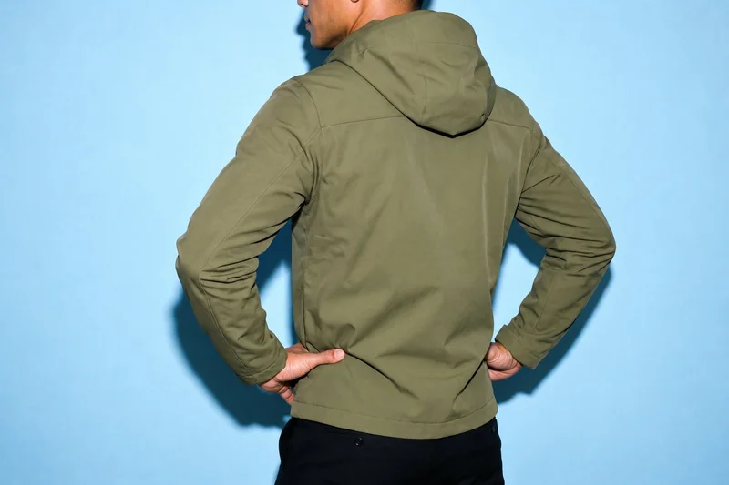 A male model is shown from the torso in an angled rear view wearing a green waterproof jacket, with his hands on his hips. An ice-blue studio wall forms the backdrop.