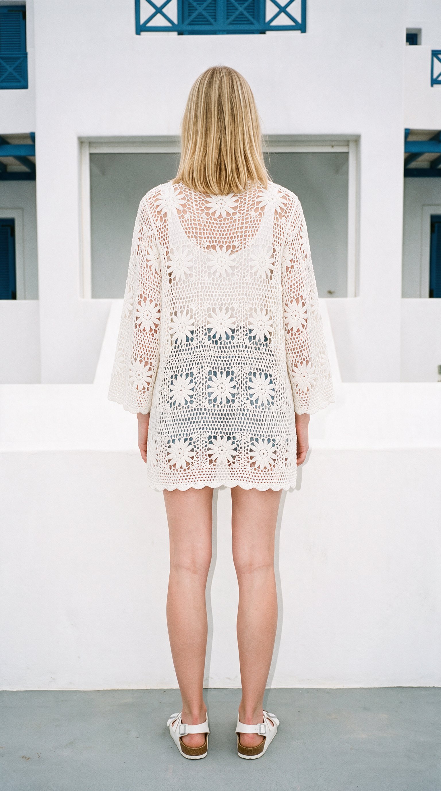A 19-year-old Caucasian woman stands straight in a full-body rear view wearing a white crochet cover-up. The image uses a white-and-blue Greek villa backdrop.