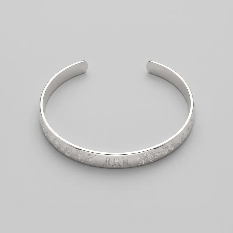 A complete women's open cuff bracelet is shown from an angled overhead view. Its continuous curved band has a visible gap between rounded blunt ends, raised polished rims along both edges, and engraved floral and leaf motifs across the front and lower side sections, with centered script initials reading “A&M.”