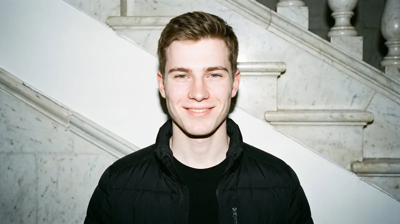 A 19-year-old Caucasian man is shown from the bust up in a front-facing view, wearing a black down jacket. His head is held level with a natural facial expression, against a marble staircase backdrop.