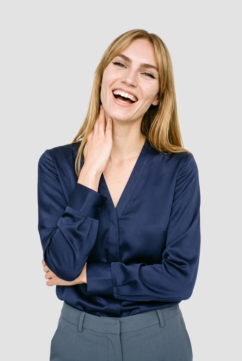 A 23-year-old Caucasian woman wears a navy satin blouse in an upper-body view from a front three-quarter angle. She is laughing with one hand on her neck, against a flat fog-grey backdrop.