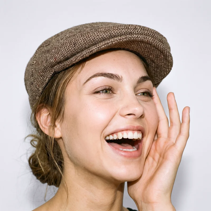A 23-year-old Caucasian woman wearing a women's brown tweed flat cap is shown in a front-view head framing with one hand at her face and a laughing expression. A porcelain studio-wall backdrop fills the background.