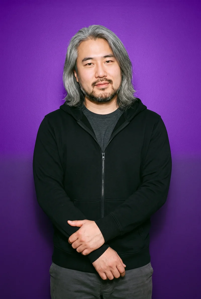 A 38-year-old Chinese man is shown from the front in an upper-body view wearing a black zip-up hoodie. His forearms are folded, and he has a curious facial expression. An indigo studio-wall backdrop fills the background.