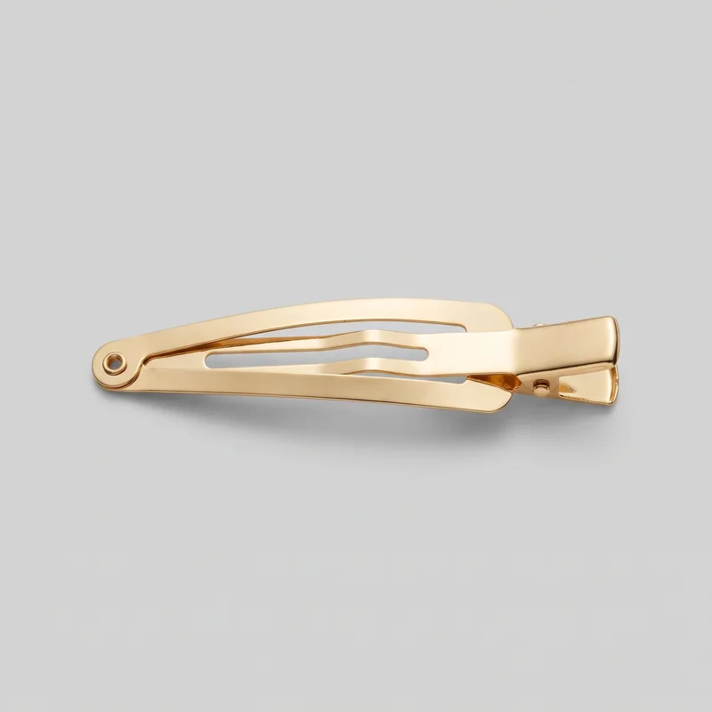 A single gold barrette-style alligator hair clip is shown as a standalone product from a side angle. Its hinged end, circular rivet opening, layered spring arms with elongated cutout slots, side rivet or peg, and flat rectangular gripping end are visible. The clip has smooth polished surfaces and rounded edges, with no visible logos, prints, patches, embroidery, or embellishments.