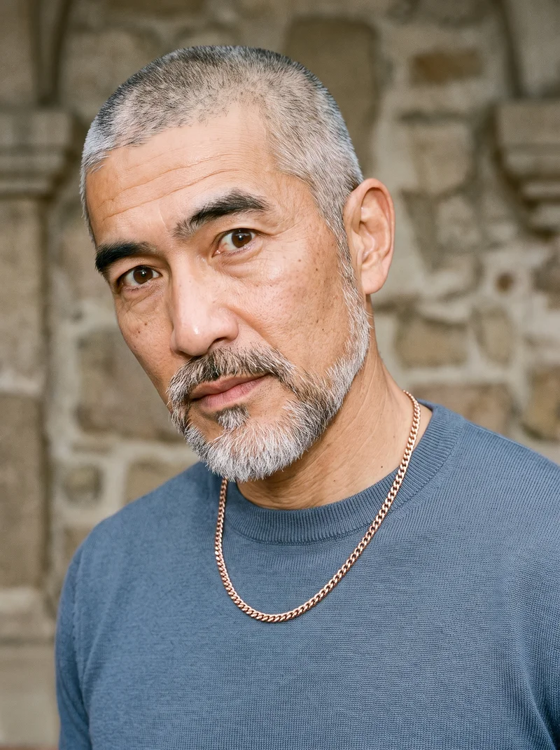 A 50-year-old Japanese man wears a men's rose gold chain in a bust composition viewed from a front three-quarter angle. His shoulder is dropped, and his curious expression is framed against a Mission Courtyard Bell Fountain backdrop.