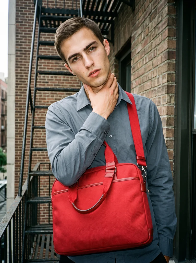 A 23-year-old Caucasian man is shown from the front in an upper-body view, wearing a red crossbody bag with one hand on his neck and a fierce expression. A fire escape serves as the backdrop.