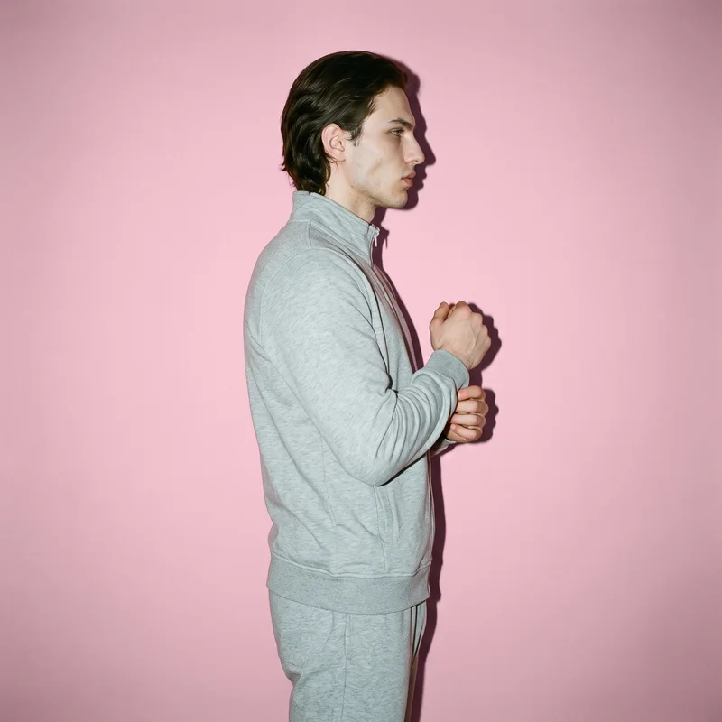 A 20-year-old Caucasian man is shown from the upper body in side view wearing a grey marl tracksuit. He has a neutral expression and is touching his wrist, with a light powder pink studio-wall backdrop.