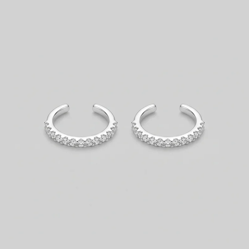 A matched pair of open C-shaped cuff hoop earrings is laid side by side. Each earring has a smooth rounded upper band and a lower arc decorated with a continuous row of round faceted stones in raised scalloped settings with fine pavé-like texture and bead-like prong details. The earrings have blunt rounded open ends and no visible posts, hooks, clasps, logos, or printed markings.