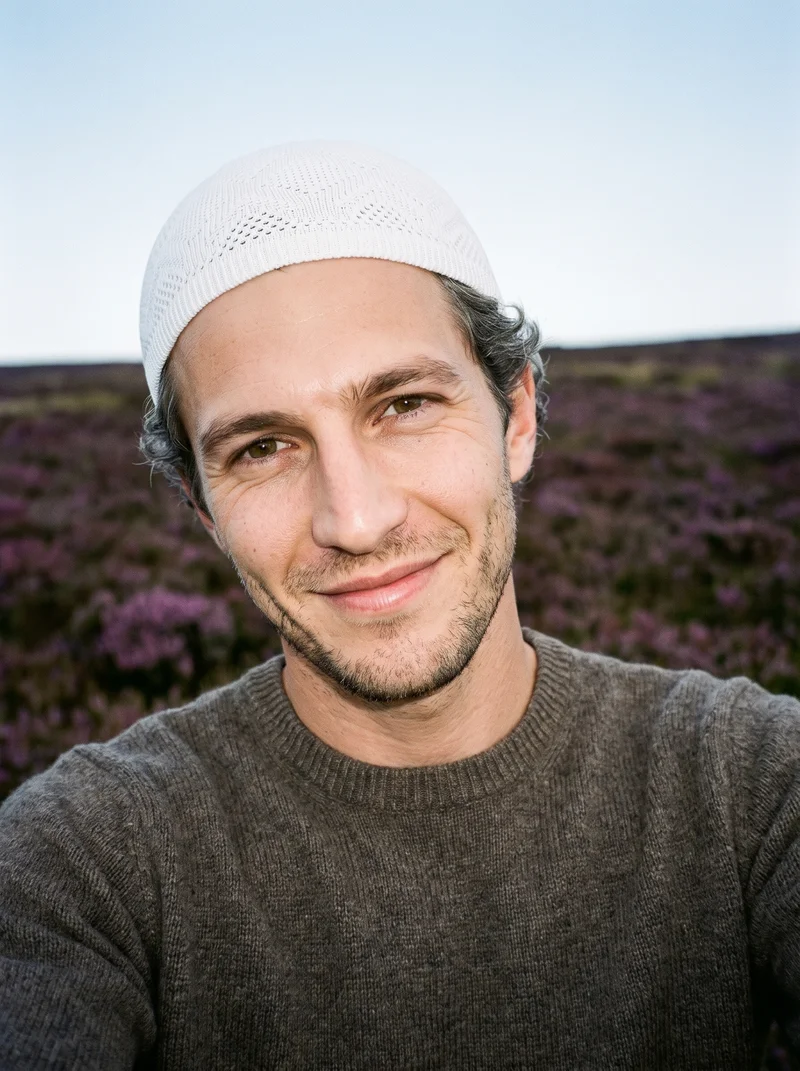 A 31-year-old Caucasian man wears a white knit kufi in a front-facing selfie with his head tilted and a smile. A Scottish Highlands moor serves as the backdrop.