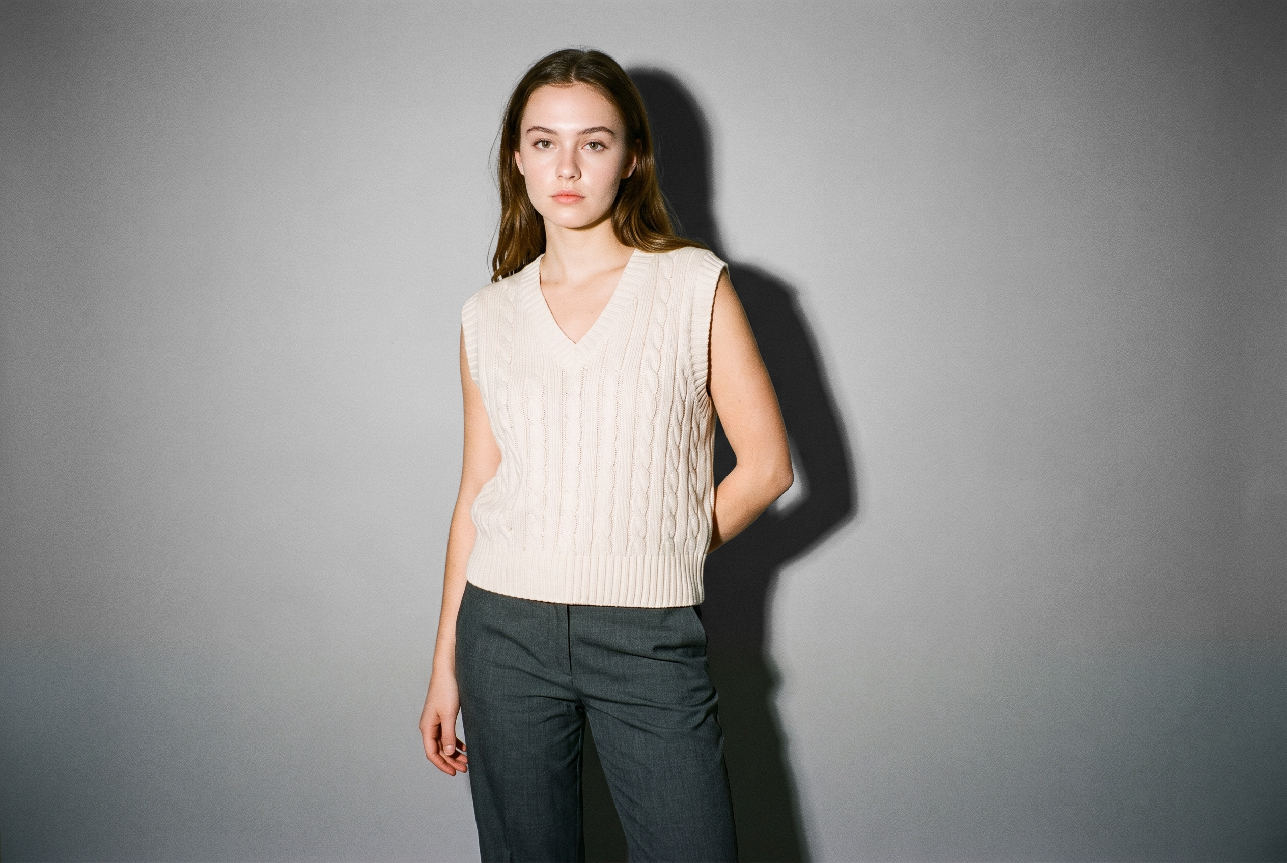 A 19-year-old Caucasian woman wears a women's cream cable-knit sweater vest in a front-facing, three-quarter-length view. She stands with her hands behind her back and a confident facial expression against a pebble grey studio-wall backdrop.