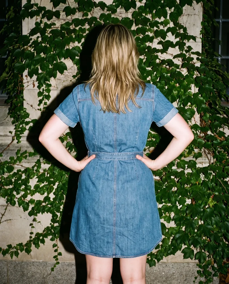 An 18-year-old Caucasian woman wears a denim shirt dress, shown from the rear in a three-quarter-length frame with her hands on her hips. An ivy-covered building forms the backdrop.