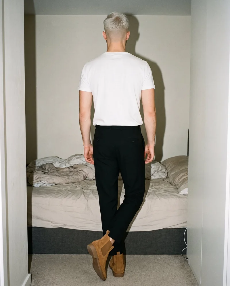 An 18-year-old Caucasian man is shown full body from the rear with his ankles crossed, wearing men’s tan suede Chelsea boots. A bedroom backdrop fills the setting.