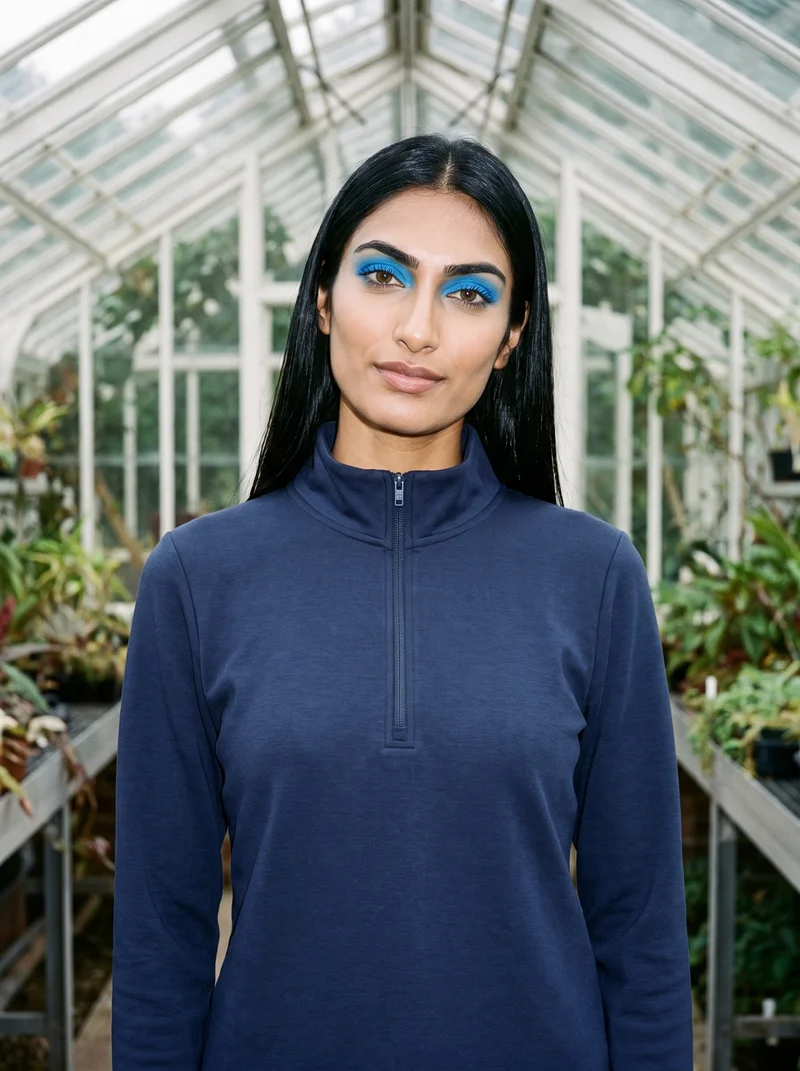 A 22-year-old Indian woman wears a women's navy quarter-zip, shown from the front in an upper-body frame with a shoulder-drop pose and natural expression. A greenhouse conservatory serves as the backdrop.