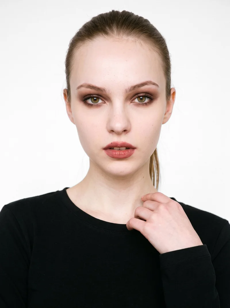A 21-year-old Caucasian woman wears a black long-sleeve tee in a front-facing bust composition, with one hand at her collarbone and a neutral expression. The garment is shown against a pure white backdrop.