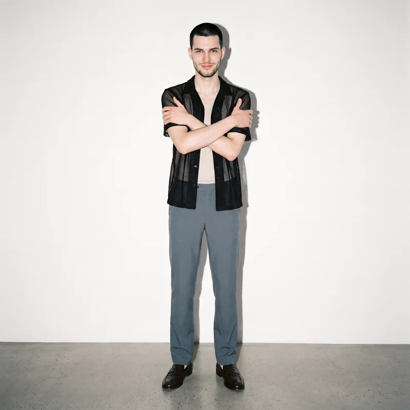 An 18-year-old Caucasian man is shown full body from the front, smiling and holding himself in a self-hug pose while wearing a black mesh cover-up. A Chantilly Lace studio-wall backdrop fills the background.