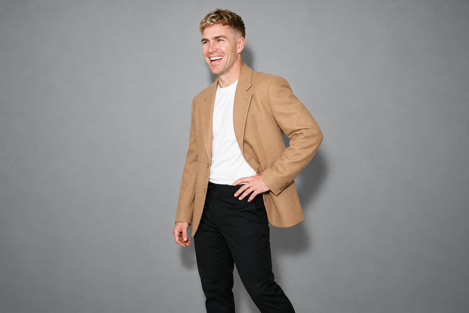 A 40-year-old Caucasian man wears a camel oversized blazer in a three-quarter-length view, angled toward the front with one hand on his hip. He is laughing against a heather grey studio-wall backdrop.