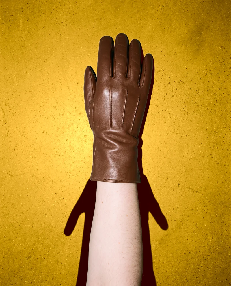 A top view shows a woman's hand and wrist wearing brown leather gloves, resting against a goldenrod studio-wall backdrop.