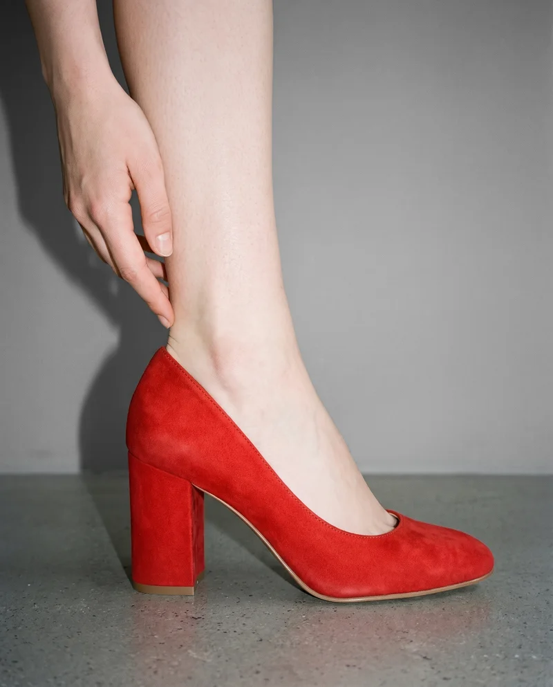 A female model wears red block heels, shown from the ankle in side view with a hand at the ankle. The shoes are presented against a pebble grey studio-wall backdrop.