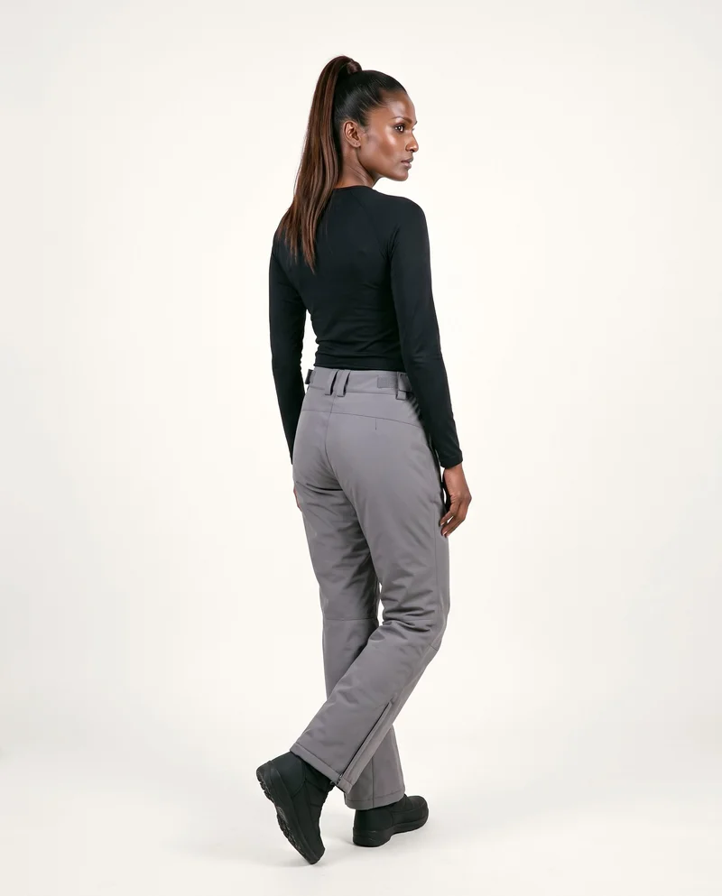 A 37-year-old Indian woman wearing women's grey waterproof ski trousers is shown full body from an angled rear view with her ankles crossed. The trousers are set against a soft ivory flat-color backdrop.
