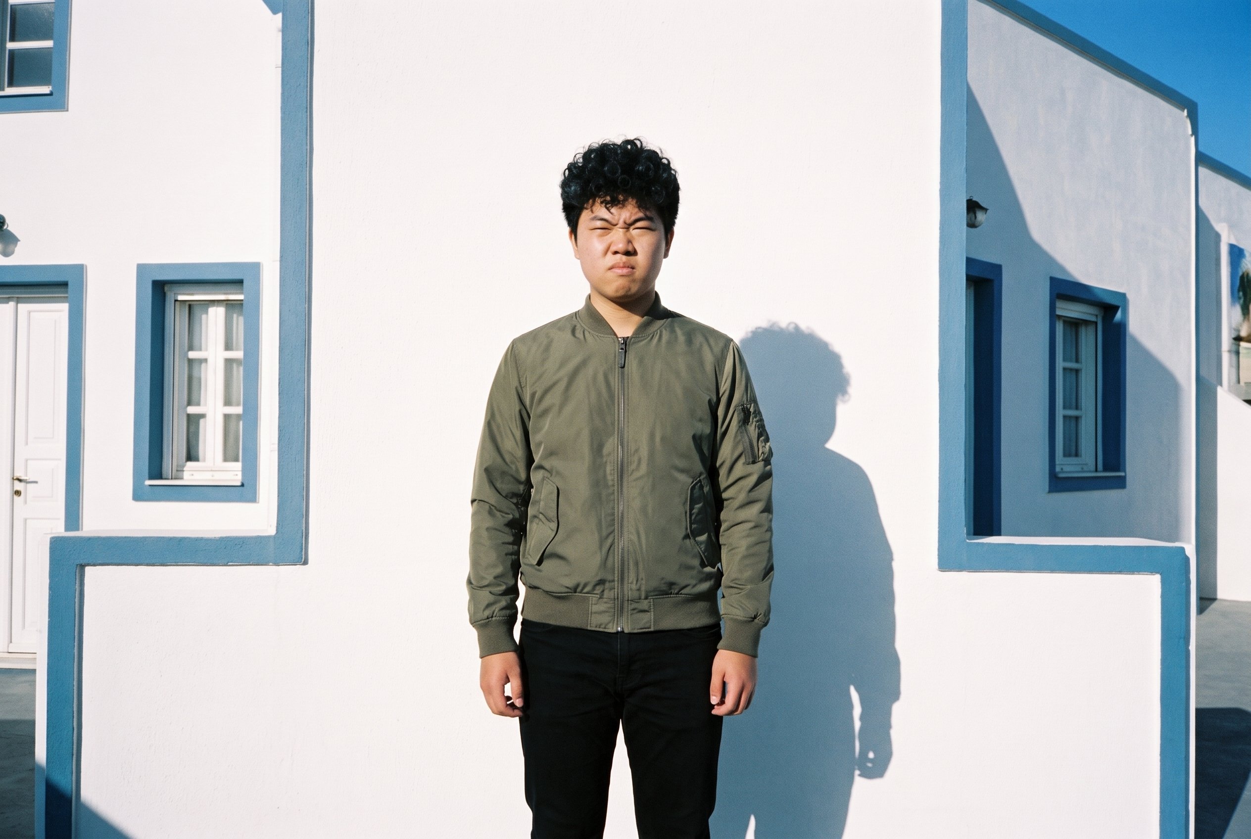An 18-year-old Chinese male model stands straight in a front-facing, three-quarter-length view wearing an olive MA-1 bomber jacket. His intense expression is visible against a Greek Villa White Blue backdrop.