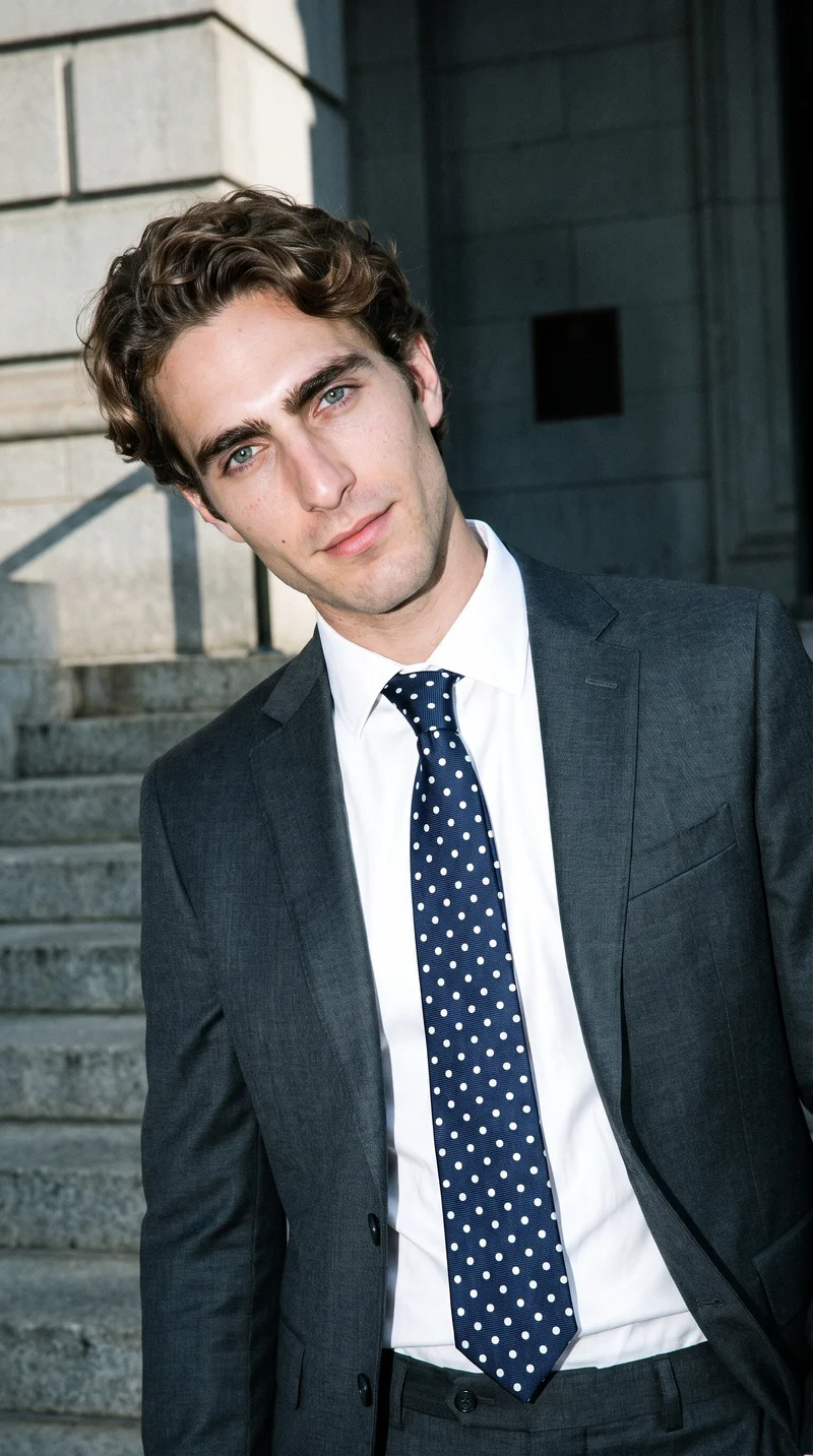 A 28-year-old Caucasian man wears a polka-dot tie in an upper-body, front-view photograph. He is shown with a natural expression and a shoulder-drop pose against a courthouse steps backdrop.