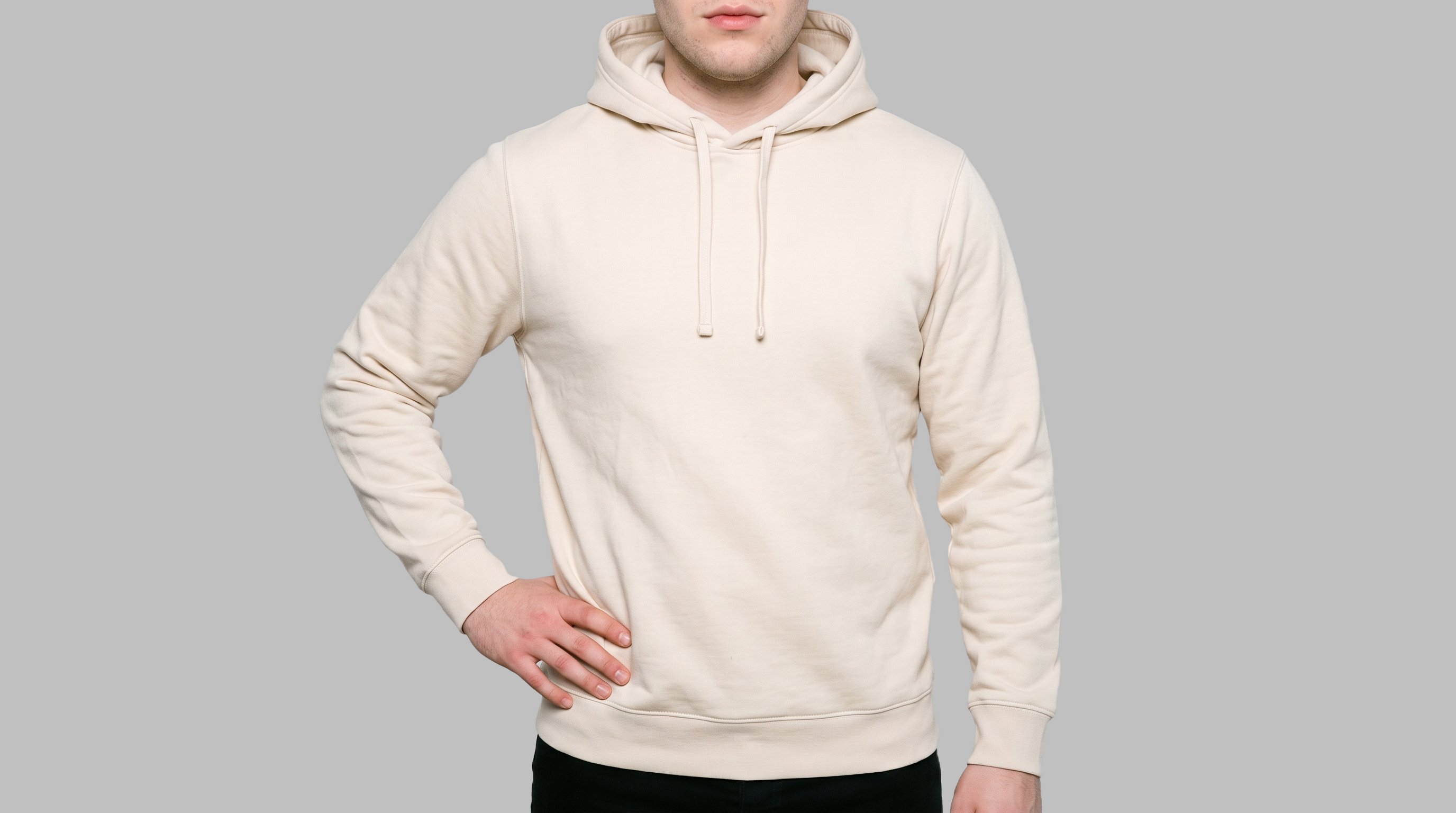 A male model is shown from the torso in a front view wearing a cream pullover hoodie, with one hand resting on his hip. The garment is presented against a flat silver mist backdrop.