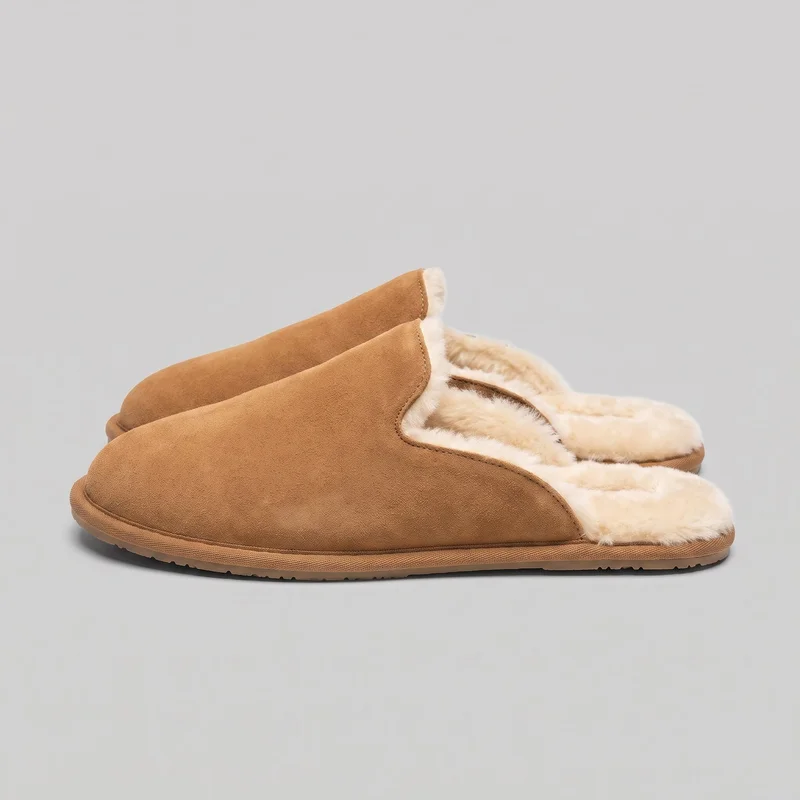 A pair of tan suede closed-toe mule slippers is shown in a side-oriented view, with one slipper mostly unobstructed and the second partially behind it. The rounded toes, open heels, exposed fuzzy lining and footbeds, stitched edges, low flat profiles, and textured treaded soles are visible.