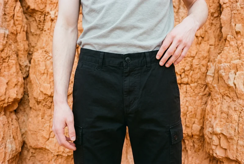 Men's black cargo pants shown in a front view, cropped at the waistline, with the model in a waist-twist pose. The Bryce Canyon Hoodoos serve as the backdrop.