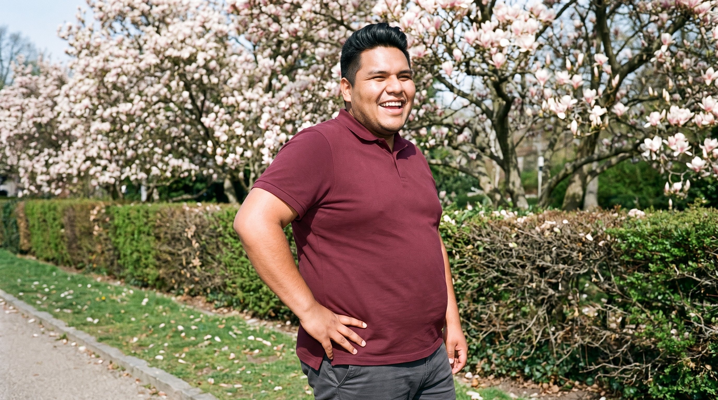 A 23-year-old Indigenous American man wears a burgundy pique polo shirt in an upper-body, angled-front view. He is laughing with one hand on his hip, against a Magnolia Tree Park backdrop.