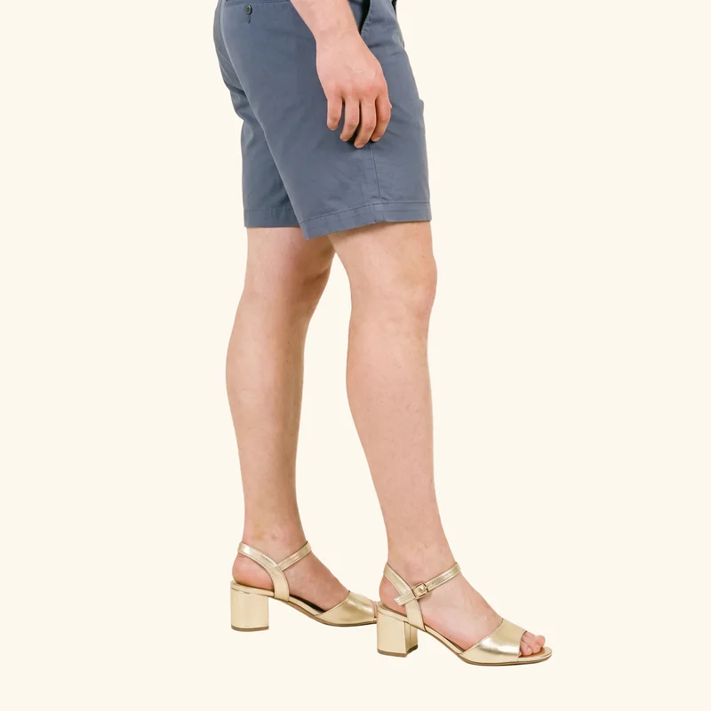 A male model's lower body is shown in side view wearing men's gold heeled sandals, with his weight resting toward the back. The sandals are presented against a creamy white flat-color backdrop.