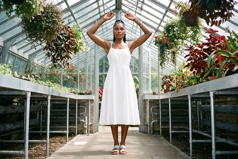A 38-year-old African woman wears a white linen midi dress in a front-facing environmental view, standing with her arms overhead and an intense expression. A greenhouse conservatory serves as the backdrop.