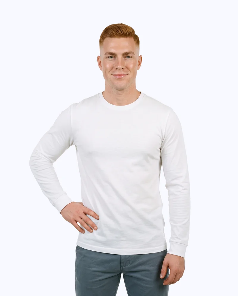 A 23-year-old Caucasian man wears a white long-sleeve tee in a front-facing three-quarter-length view. He is smiling with one hand on his waist, against a flat porcelain-colored backdrop.