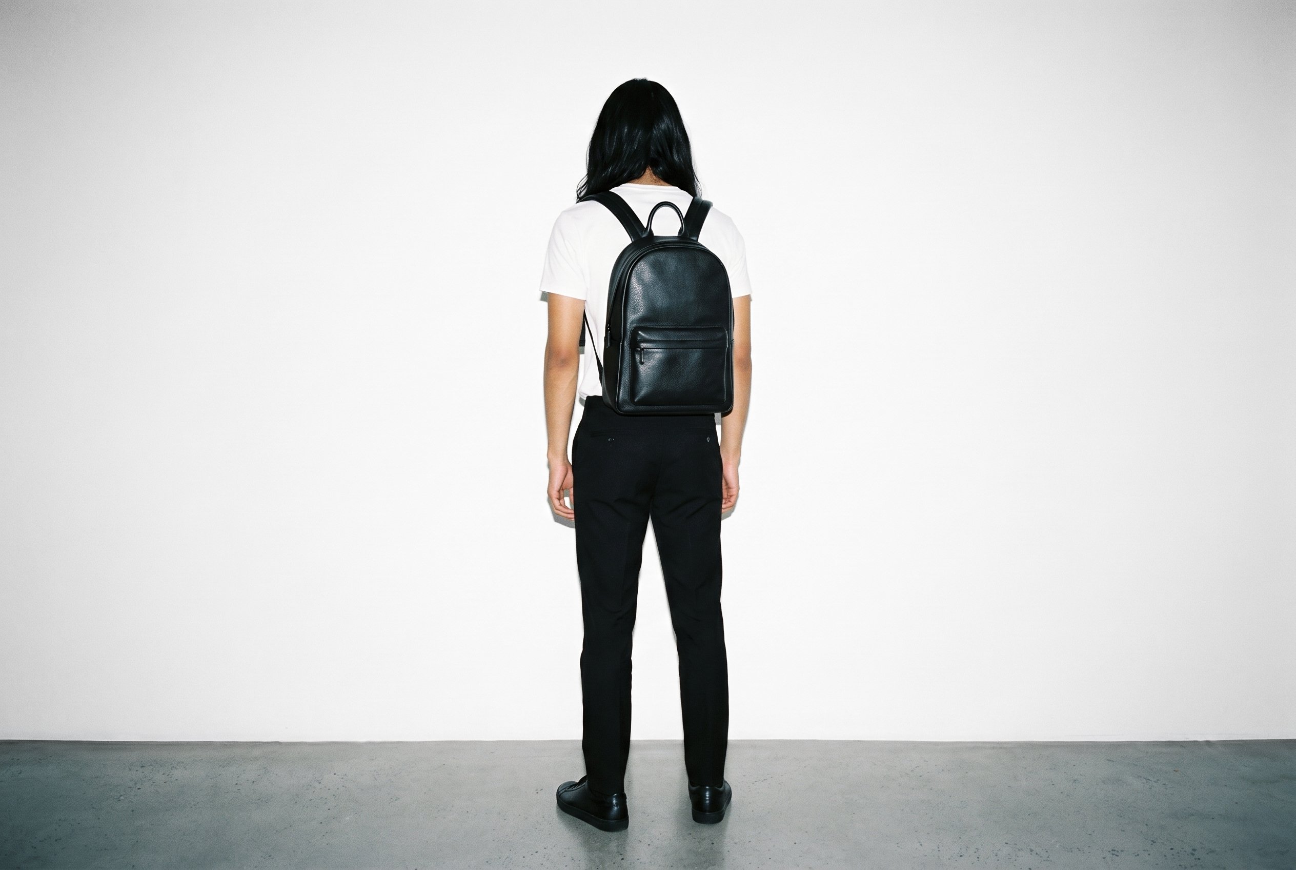 An 18-year-old Hispanic male model stands straight in a full-body rear view wearing a black leather backpack. The product is shown against a pure white studio-wall backdrop.