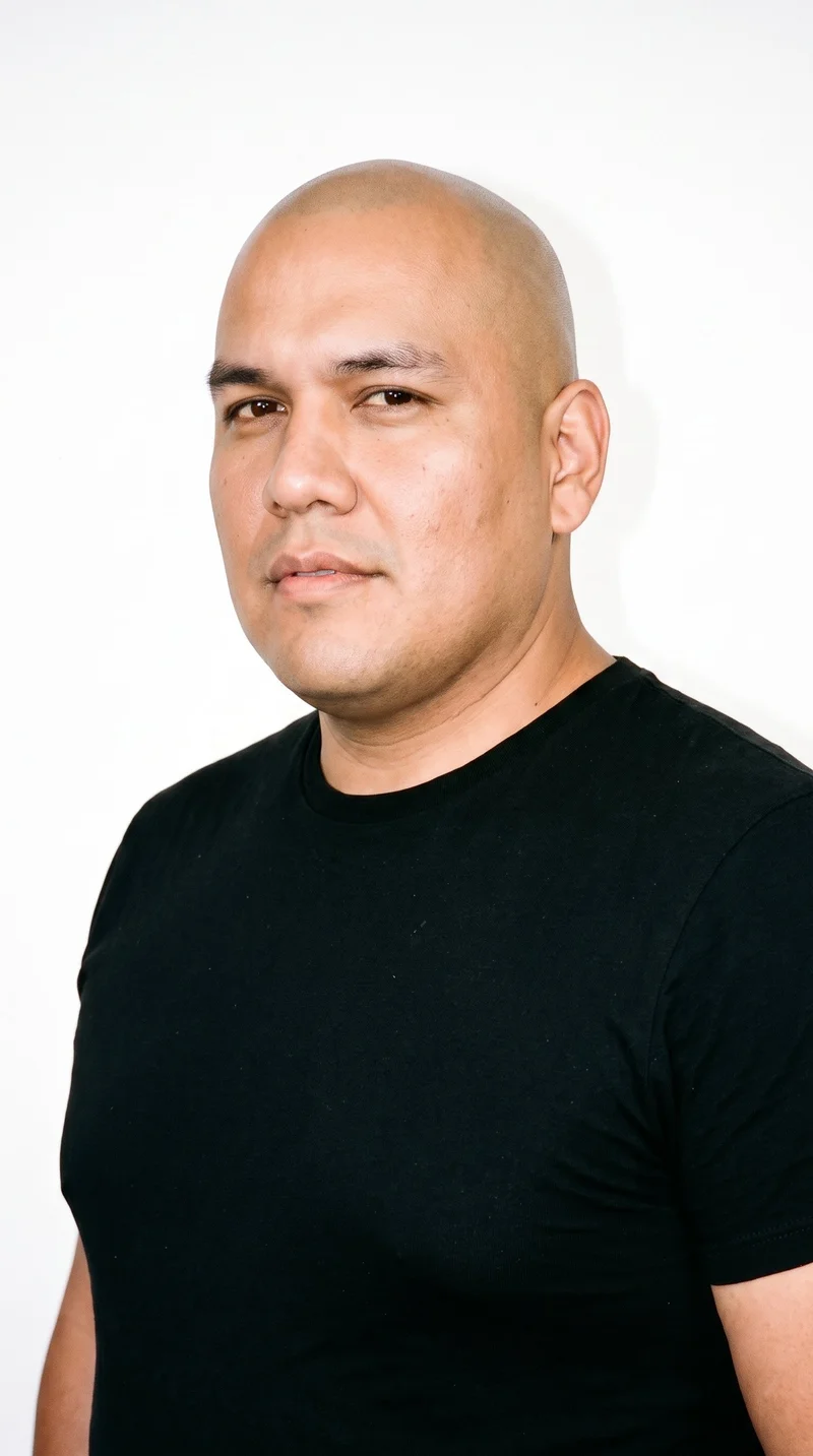 A 40-year-old Indigenous American man with a medium, evenly warm golden skin tone and a rugged face defined by weathered, characterful features and a heavy jaw. He has medium-thick eyebrows, very dark near-black eyes, and medium lips. His scalp is cleanly and uniformly bald, with no facial hair and a smooth face.