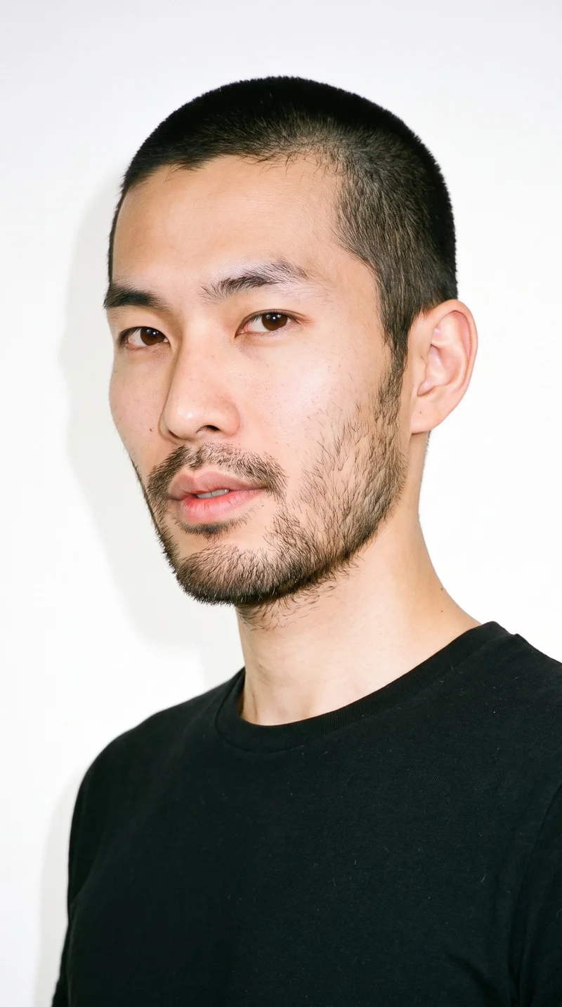 A 32-year-old Japanese man with a light porcelain skin tone and neutral undertone has a Roman face shape, marked by strong classical features, a prominent nose line, and a defined jaw. His thick eyebrows frame very dark, near-black eyes, and he has full lips. His black hair is clipped into a very short, even buzz cut, and his face has short, even three-day beard growth with a closely groomed texture.