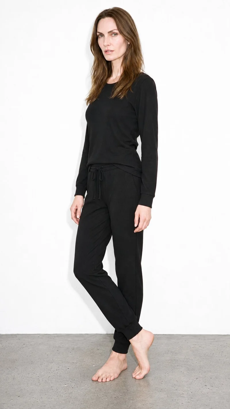 A 56-year-old Caucasian woman wears a women's black ribbed loungewear set against a pure white studio-wall backdrop. The full-body image is shown from an angled front view, with her ankles crossed and an editorial facial expression.