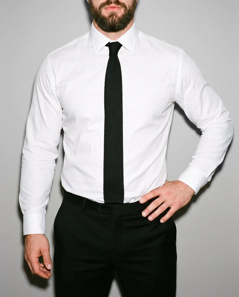 A man is shown from the torso in a front-facing view, wearing a black knitted tie with one hand on his hip. A Silver Mist studio-wall backdrop frames the composition.